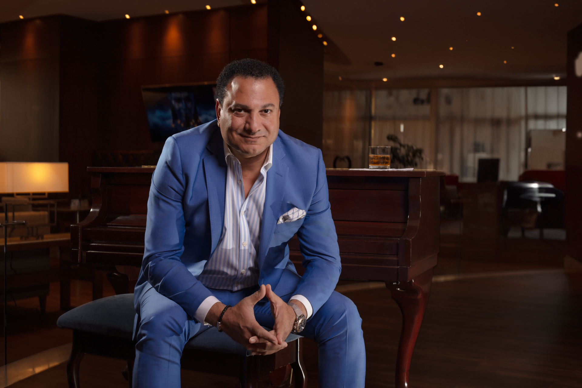 Mark Bastorous in a blue suit seated in a piano lounge, looking directly at the camera